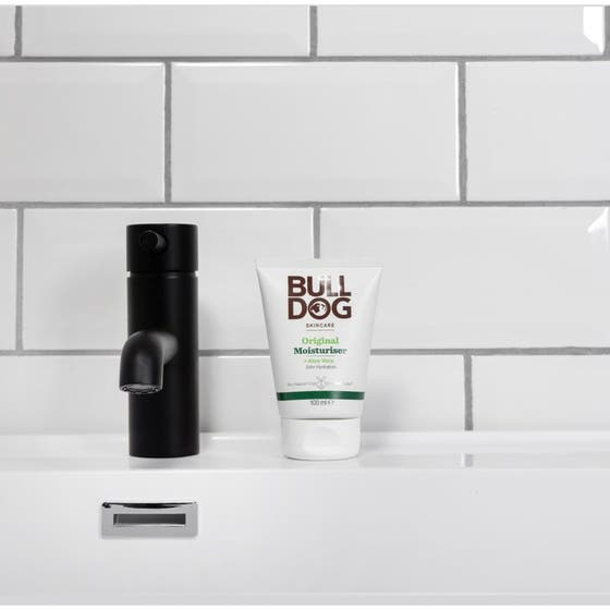 Bulldog Original Moisturiser 100 ml återfuktande ansiktskräm i vit tub bredvid en svart kran i badrumsmiljö.